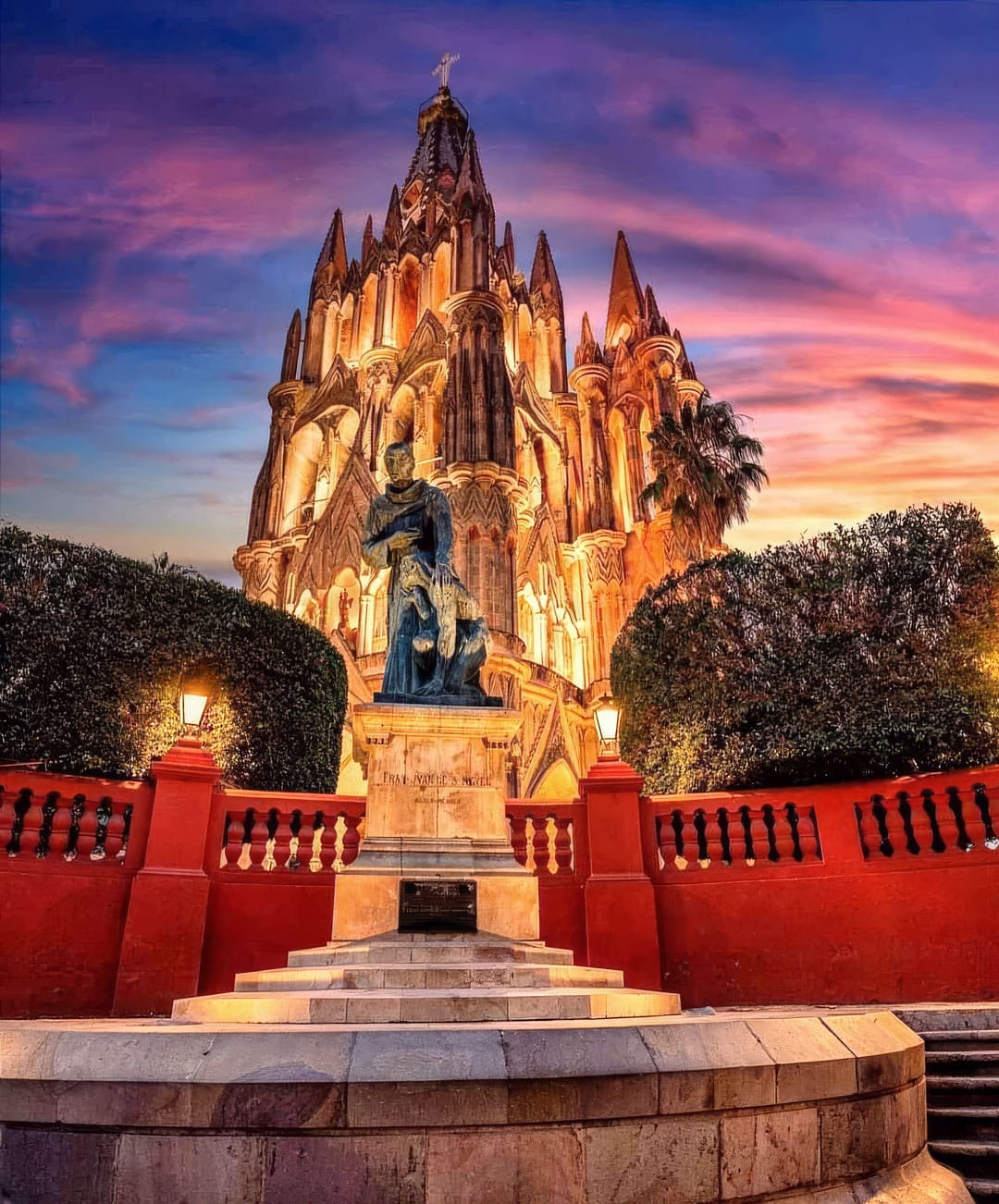 Gothic cathedral lit at sunset with a statue in a pedestal plaza in front and a red wall surrounding the area.