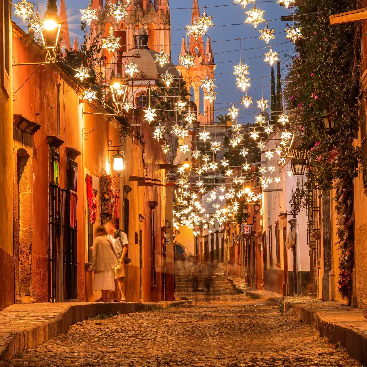 Warmly lit cobblestone street at twilight with hanging star lanterns and a church in the background.