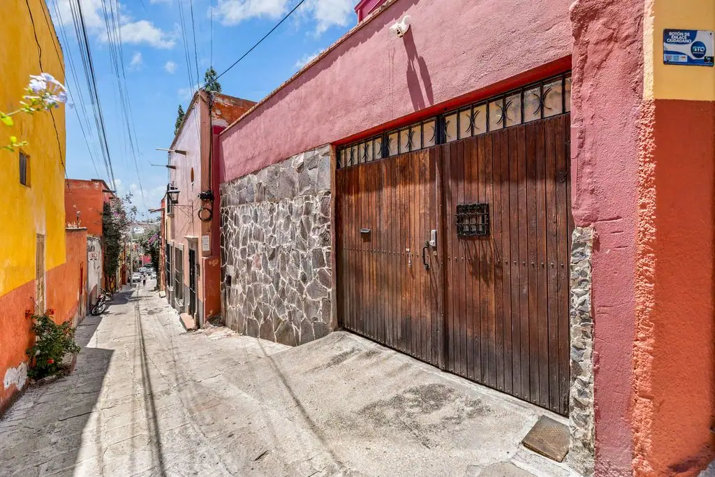 Narrow sunlit alley in a colorful neighborhood, pink and orange buildings, with a large wooden gate and stone wall, San Miguel de Allende
