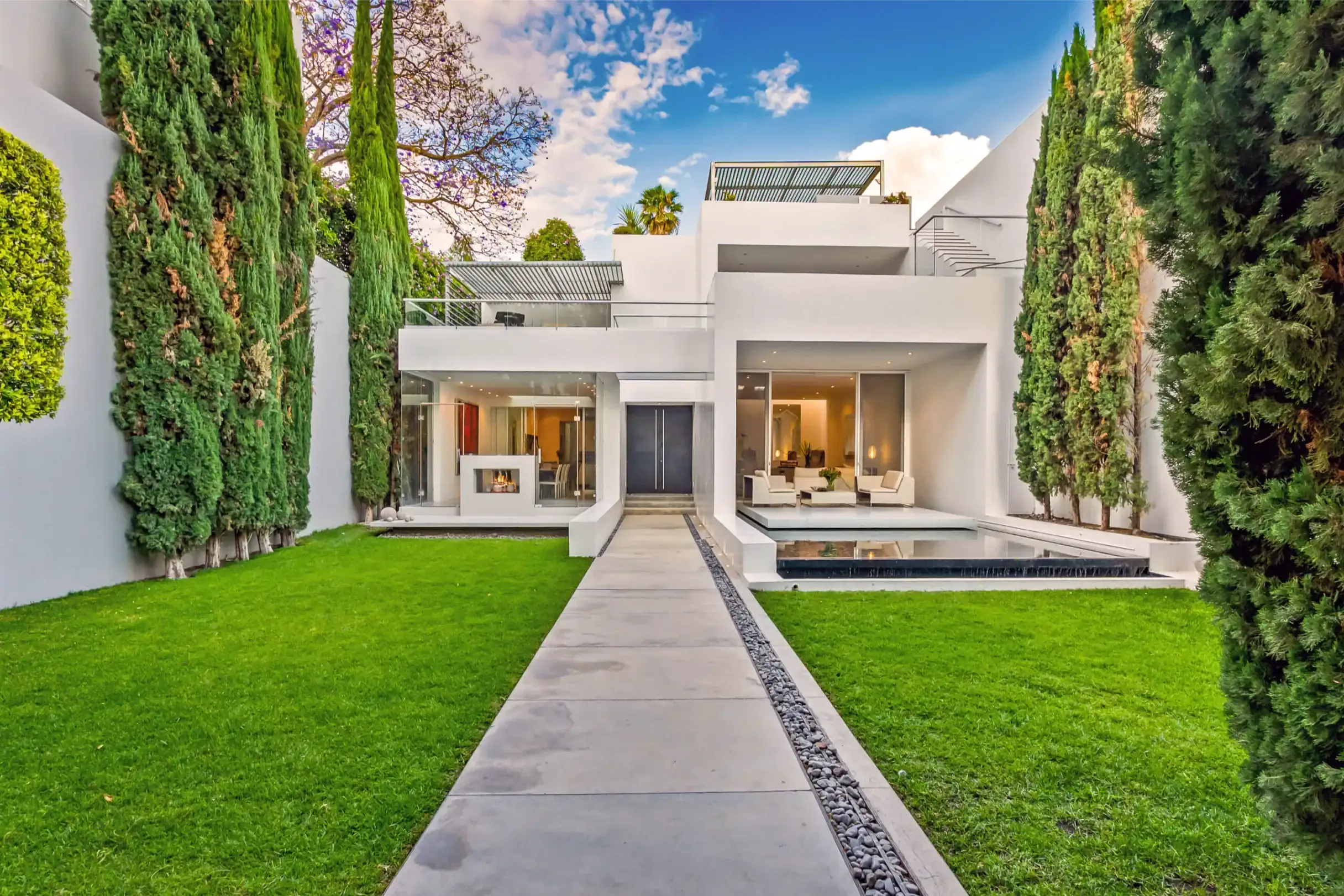 Modern white two-story house with glass doors, surrounded by green lawn and tall hedges, a walkway leads to the entrance, San Miguel de Allende