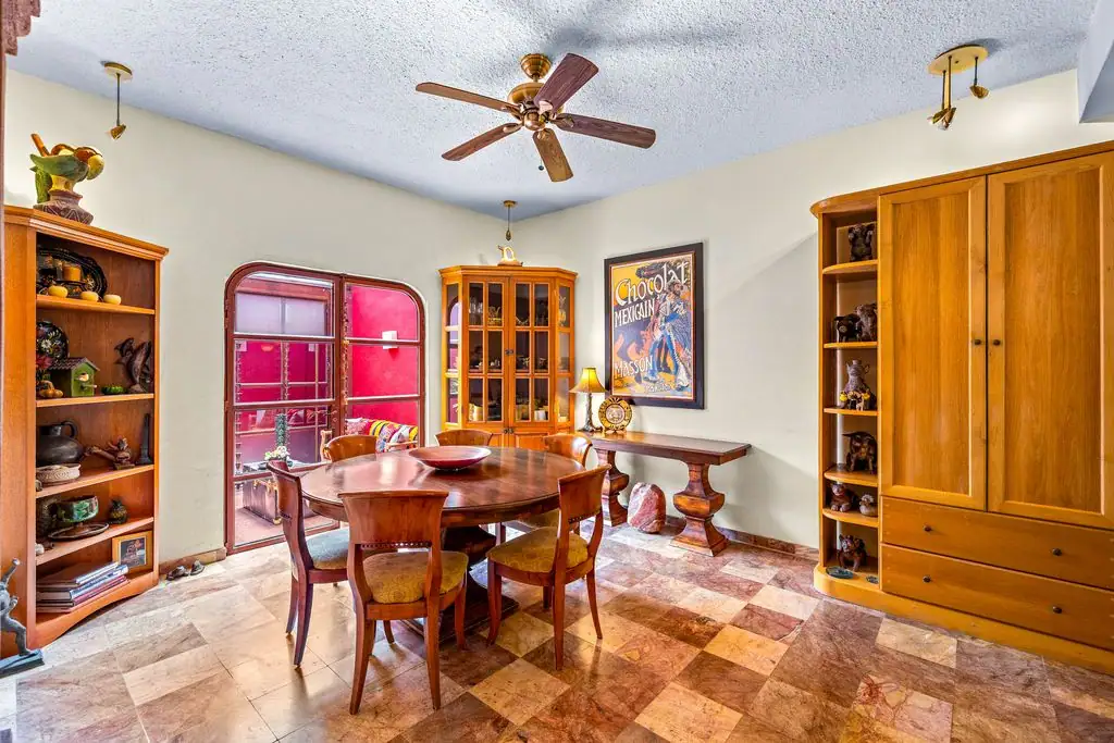 Casa Ave, dining room with a round wood table and wood chairs, a corner glass cabinet, wood bookshelves, a framed Chocolat Mexicain poster and an arched door to a red courtyard, San Miguel de Allende