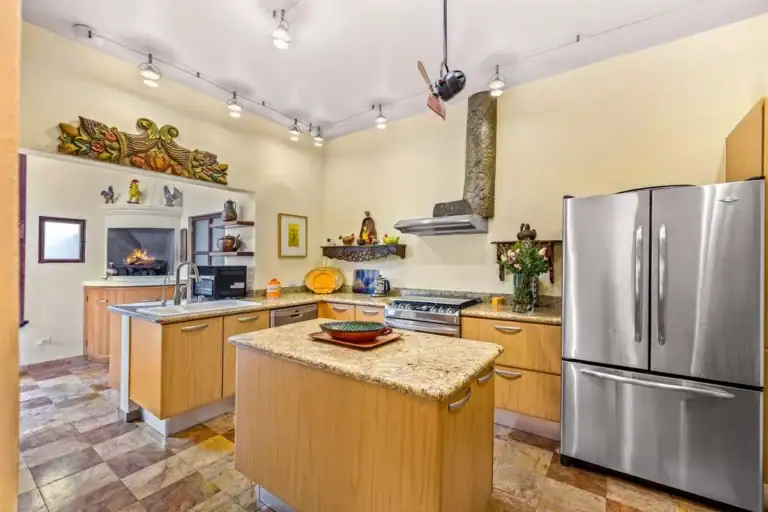 Open kitchen with a granite-topped island, light wood cabinets, stainless steel refrigerator, and range hood; fireplace area visible in adjacent room, San Miguel de Allende