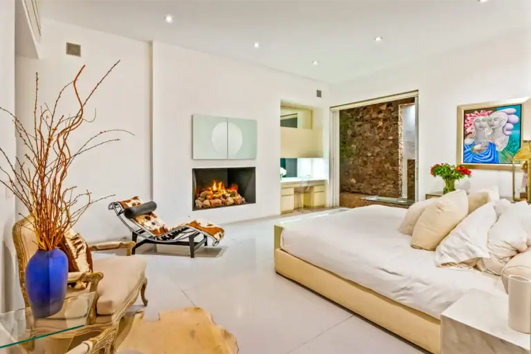 Modern living room with a lit fireplace, white walls, beige sectional couch, two-panel circular art above the fireplace, and a blue vase with dried branches on glass tables nearby, San Miguel de Allende