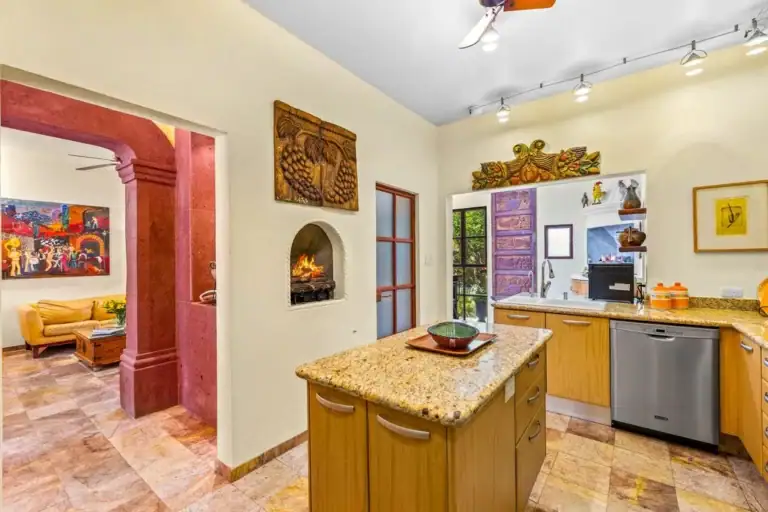 Sunny kitchen with granite island, wooden cabinets, and a stainless dishwasher; purple doorway opens to a colorful living room, San Miguel de Allende