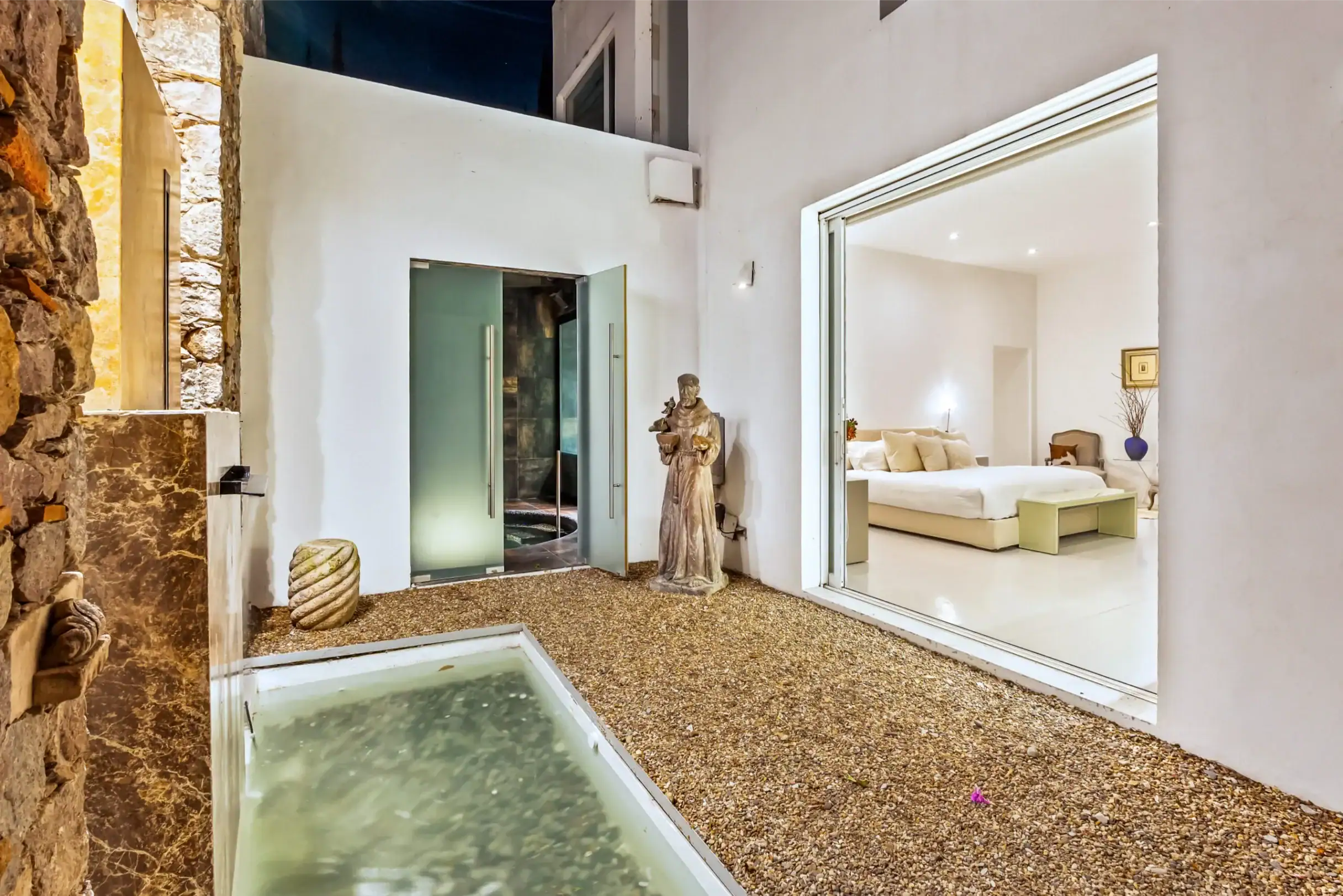 White-walled courtyard with a shallow water channel, pebbled ground, a stone wall on the left, and a statue beside glass doors opening to a bright living room, San Miguel de Allende