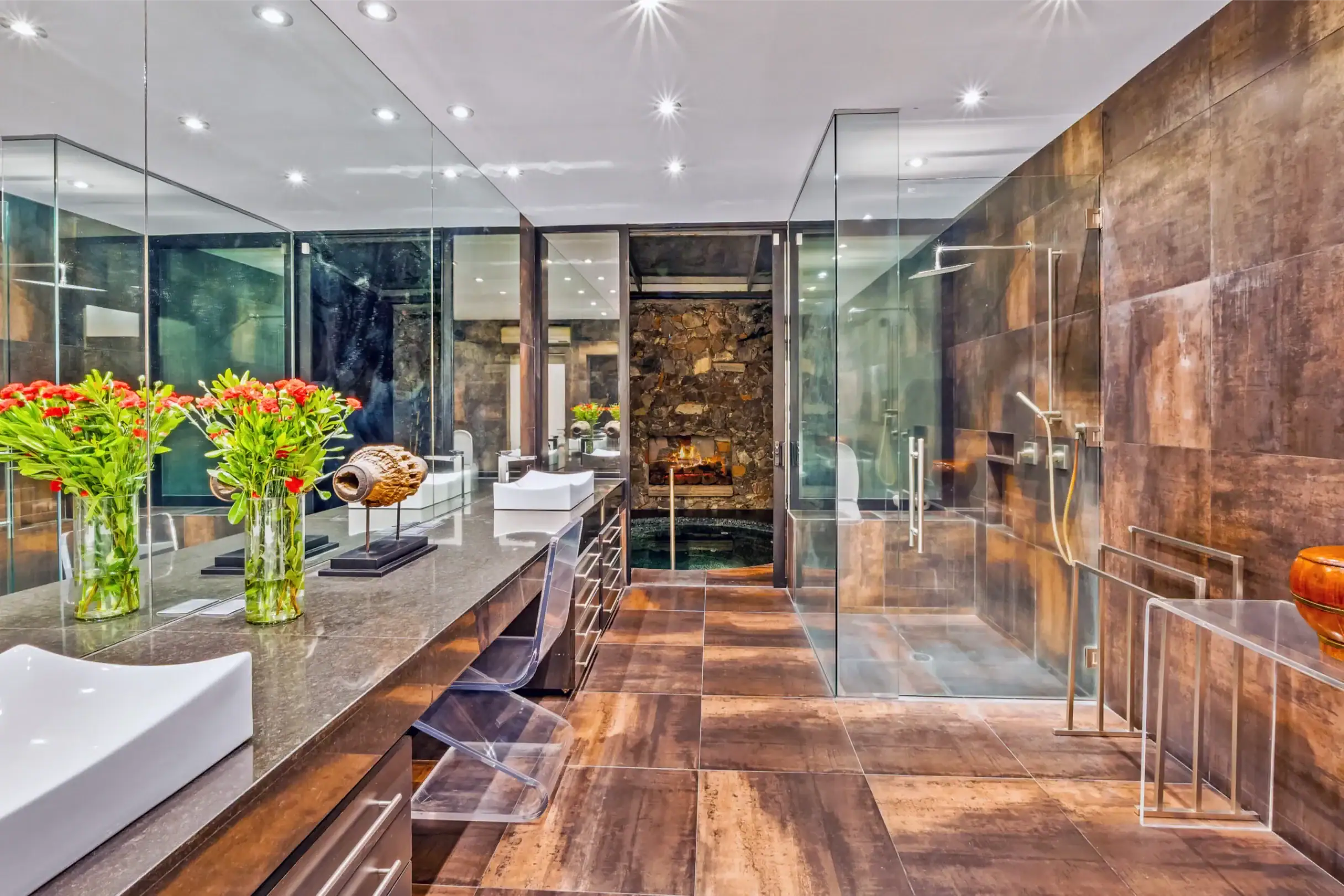 Luxurious modern bathroom showroom with glass walls, dark wood and stone tiles, and white vessel sinks along a long countertop with red flower arrangements in glass vases, San Miguel de Allende