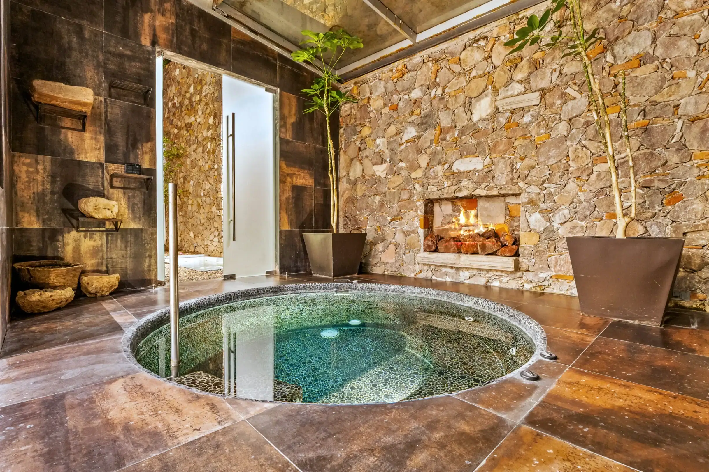 Indoor spa room with a circular mosaic hot tub, stone walls, and tall potted plants. A fireplace sits against the stone wall, San Miguel de Allende