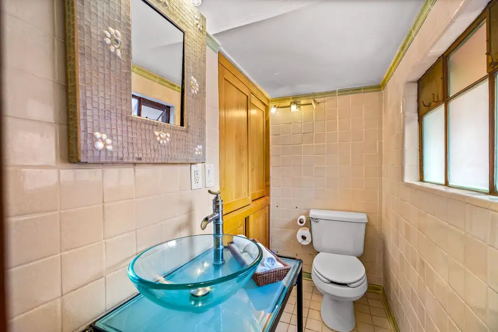 Small bathroom with a glass vessel sink on a glass counter, beige tiled walls, and a wooden storage cabinet beside a toilet, San Miguel de Allende