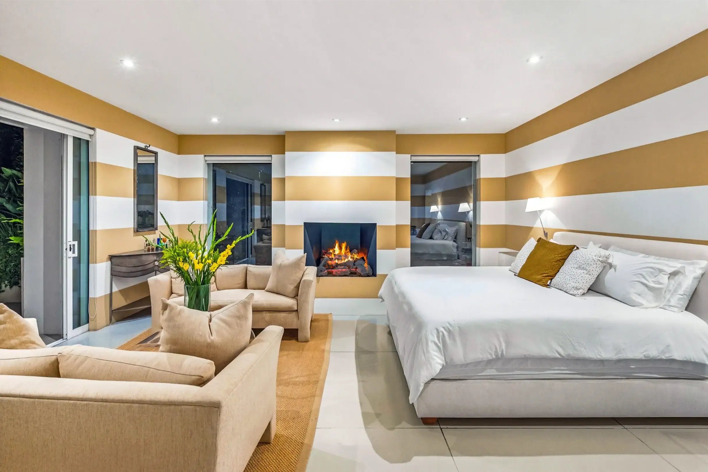 Modern hotel suite with two white beds, beige seating, and a central fireplace, against striped gold and white walls with sliding doors nearby, San Miguel de Allende
