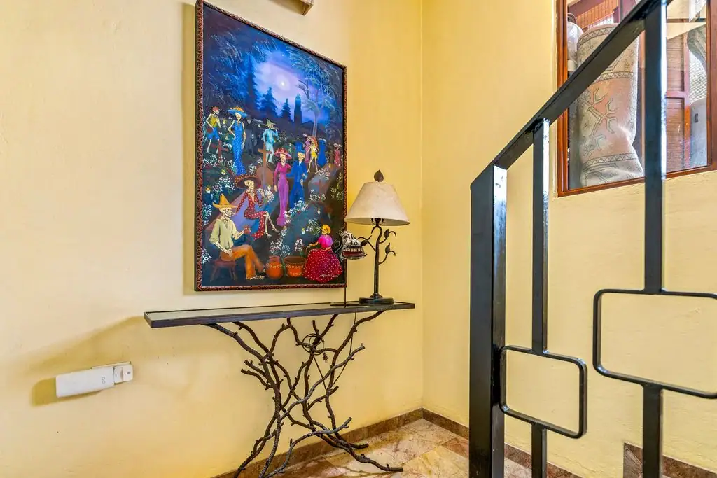 Yellow-walled stair landing with a colorful framed painting, a metal branch-table, and a table lamp near a black railing, San Miguel de Allende