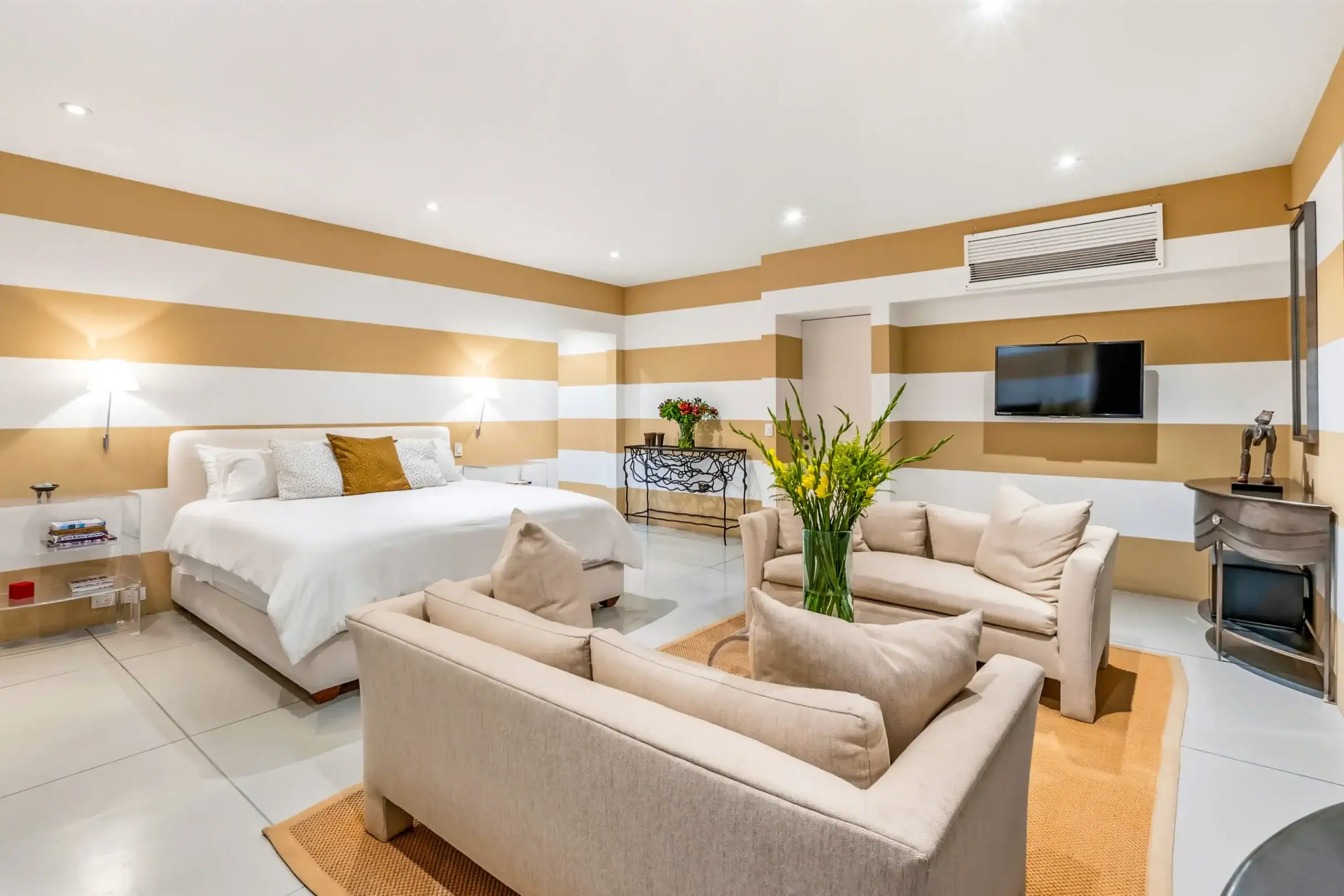 Hotel suite living area with striped tan and white walls, a white bed, beige sofas, and a wall-mounted flat-screen TV, San Miguel de Allende