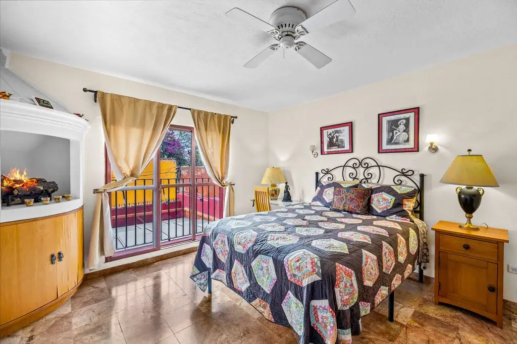 Cozy bedroom with a metal-framed bed, colorful quilt, two wooden nightstands, and beige curtains over glass balcony doors, San Miguel de Allende