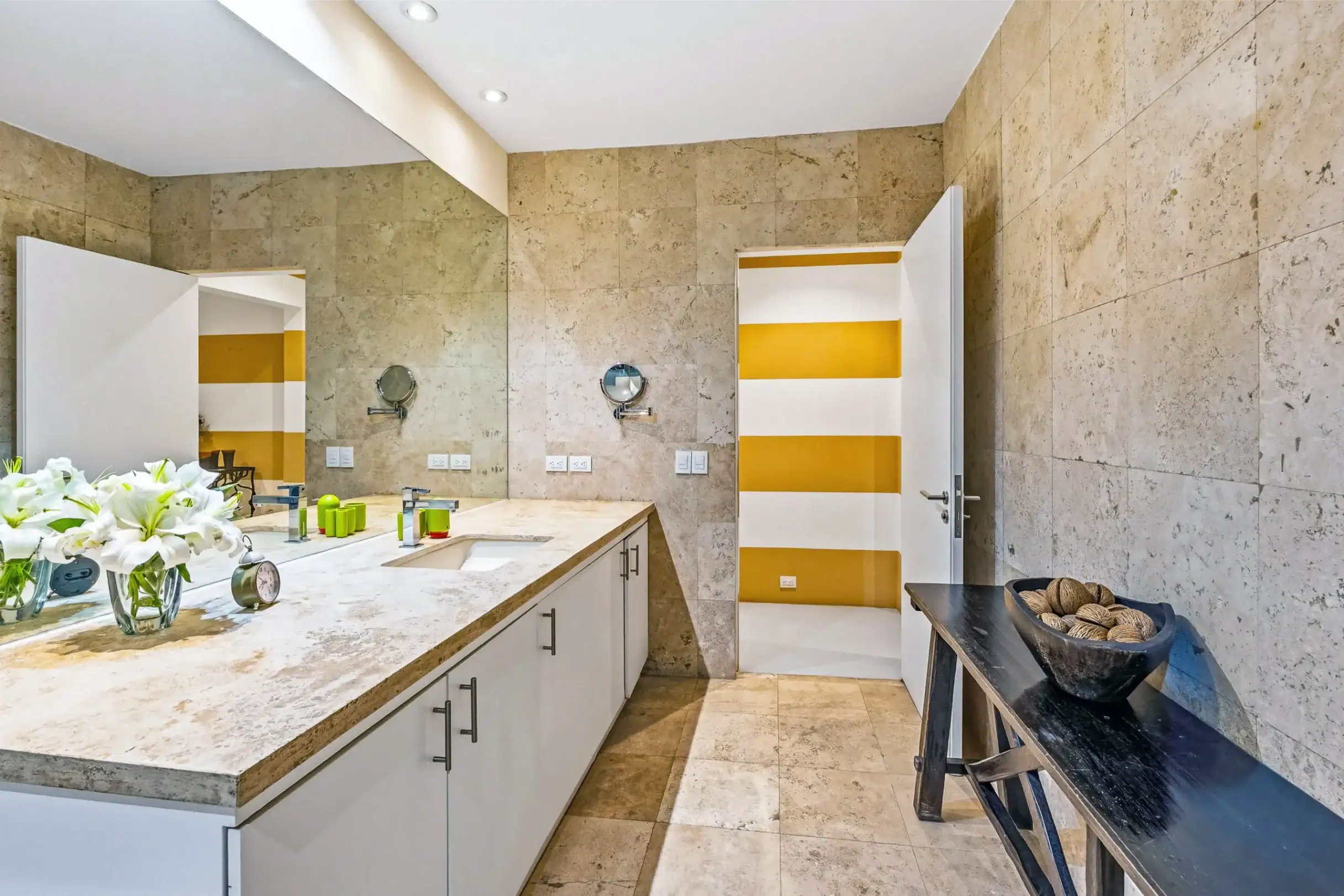 Modern bathroom vanity with marble countertop, white cabinets, beige tiled walls, and a bold yellow-and-white striped door in the background, San Miguel de Allende