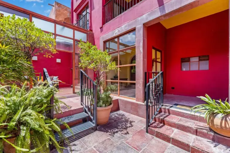 Red-walled courtyard with glass storefront, potted plants, and black metal stairs leading to doors, San Miguel de Allende