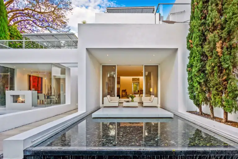 Modern white house with open living area and glass walls, seating outdoors beside a reflective pool and surrounding greenery on a sunny day, San Miguel de Allende