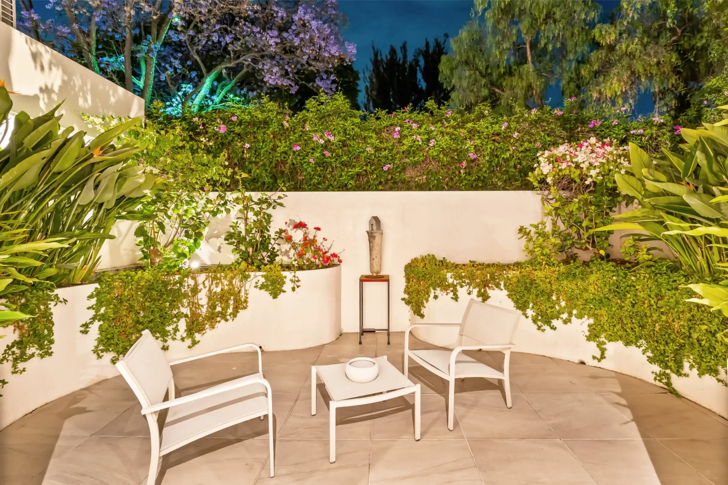 Outdoor patio with white metal chairs around a small table, lush green hedges and pink flowers, nighttime lighting in a tranquil garden setting, San Miguel de Allende