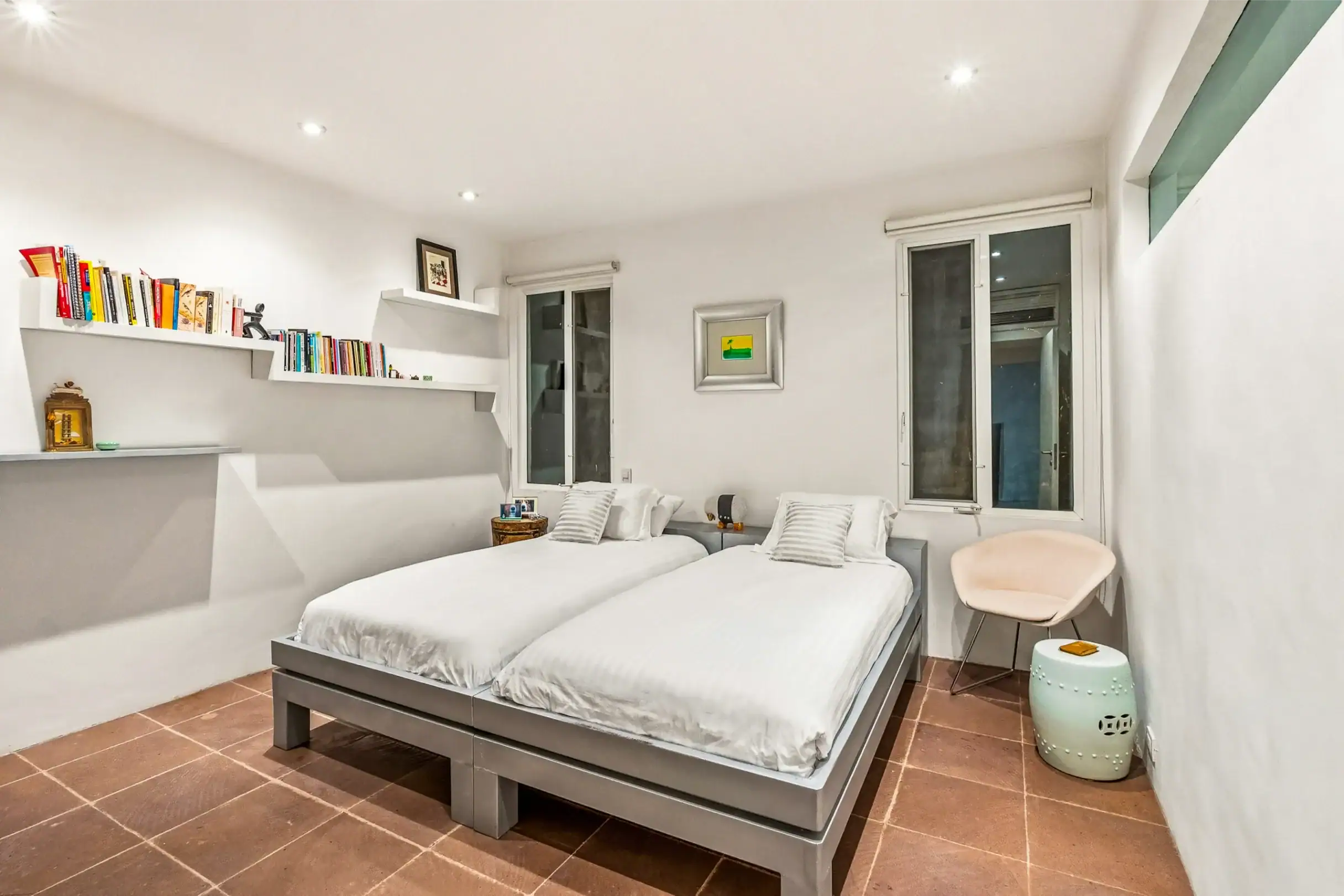 Bright modern bedroom with two white-topped twin beds, wall-mounted shelves with books, and a mint-green stool in the foreground, San Miguel de Allende