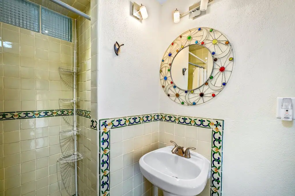 Small bathroom with a round decorative wire-frame mirror above a white oval sink, colorful tile border, and a tiled shower with a wire rack shelf, San Miguel de Allende