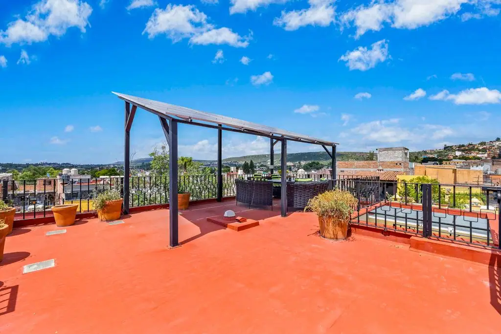 Rooftop terrace with a red concrete deck, black pergola, wicker seating, and potted plants overlooking a distant city skyline under a blue sky, San Miguel de Allende
