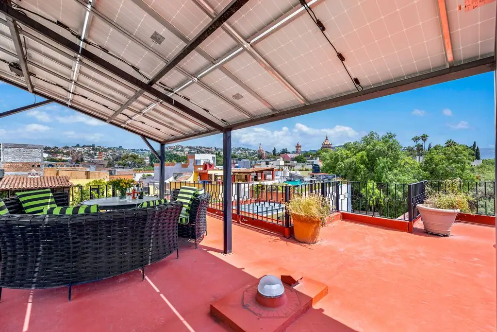 Rooftop terrace with circular black wicker seating and green cushions under a solar-panel canopy, red floor, railing, and potted plants overlooking a city skyline, San Miguel de Allende