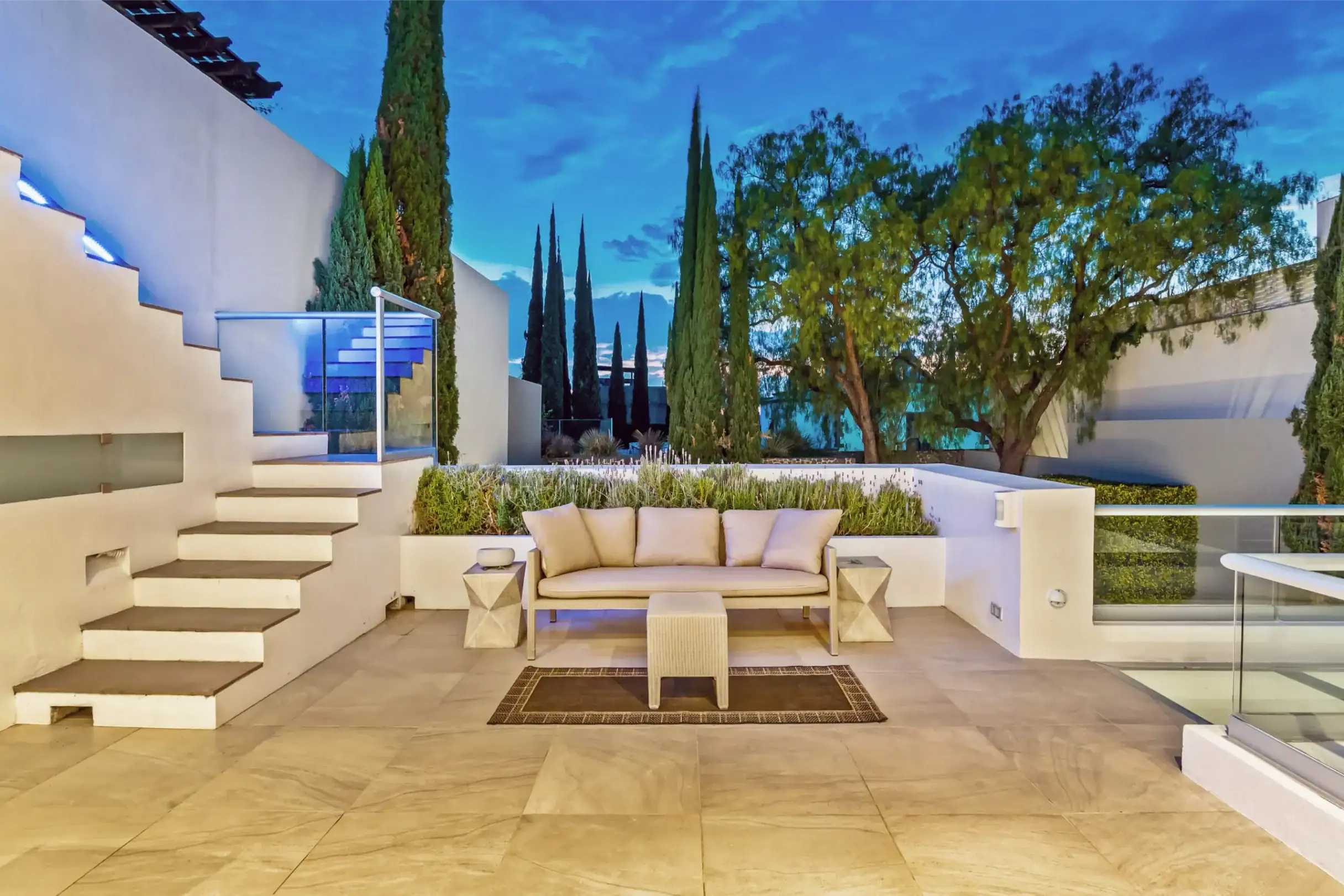 Modern outdoor terrace with white walls, steps, glass railing, and a beige sofa under a blue evening sky with tall cypress trees, San Miguel de Allende