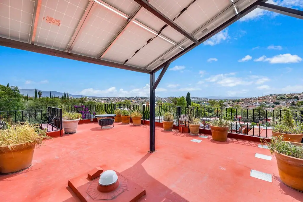 Rooftop terrace with red floor, large potted plants, and a black railing overlooking a city and hills under a blue sky, San Miguel de Allende