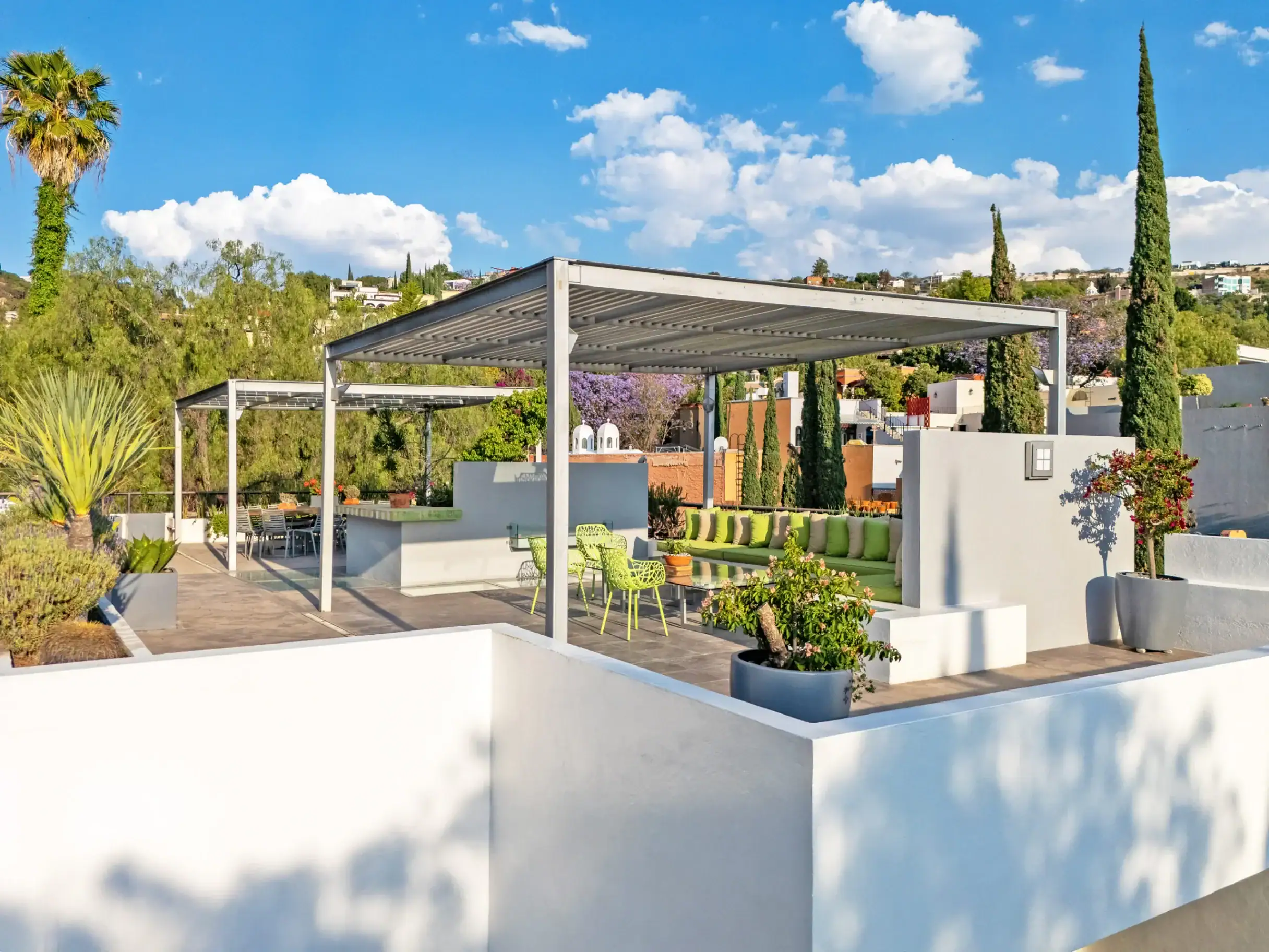 Outdoor rooftop terrace with a metal pergola, lime-green chairs, and white walls, surrounded by potted plants under a bright blue sky, San Miguel de Allende