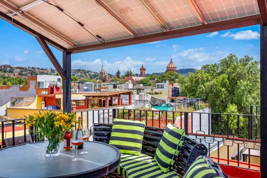 Balcony seating area with a round table, flowers, and striped green cushions, overlooking colorful rooftops and a domed building in the distance, San Miguel de Allende