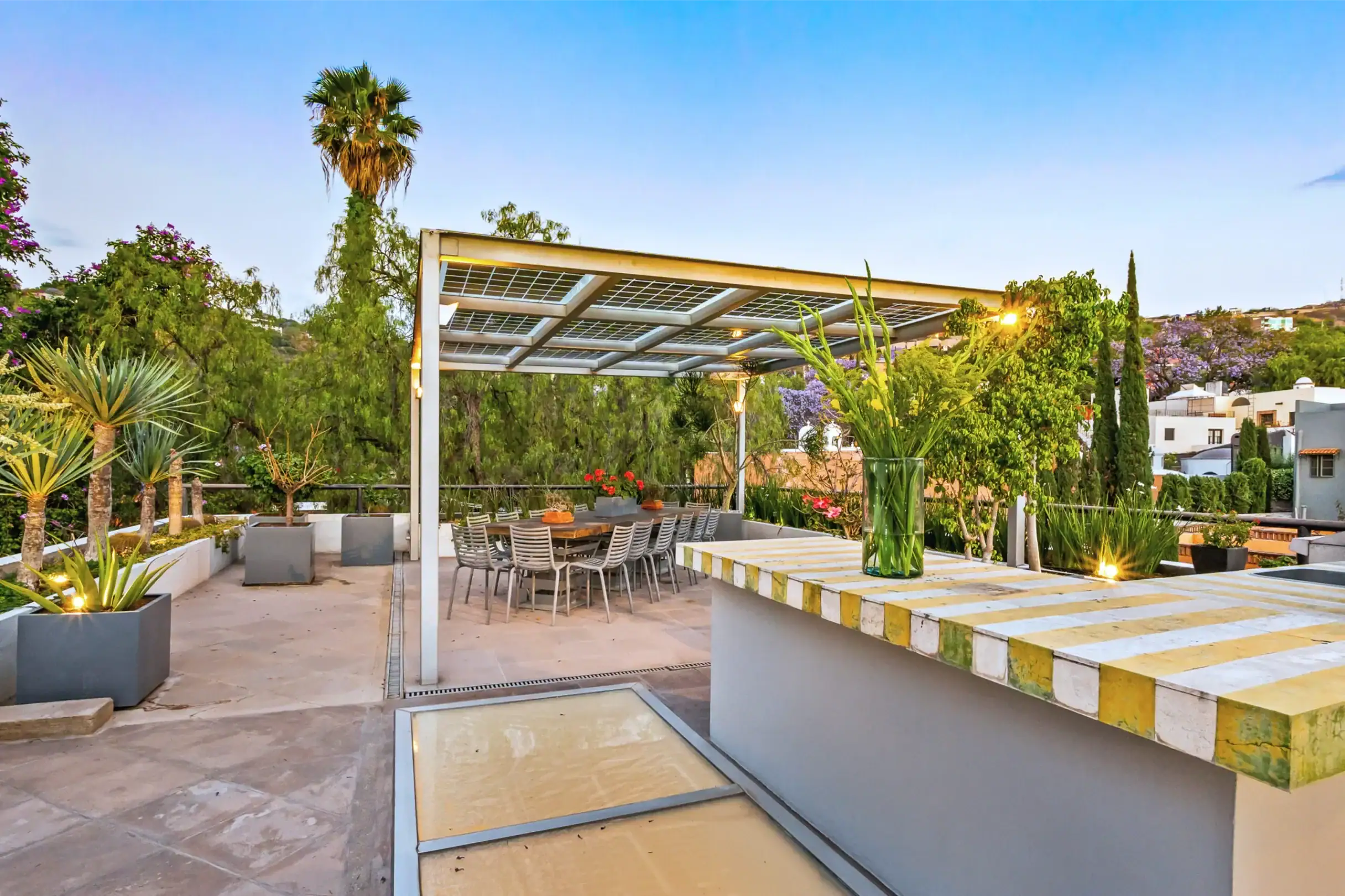 Outdoor patio with a long dining table under a metal pergola, surrounded by potted plants and greenery at dusk/light, San Miguel de Allende