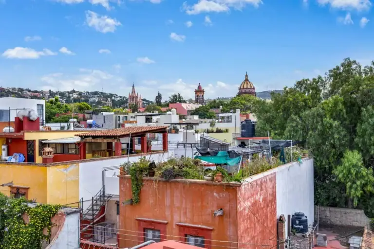Colorful rooftops and terraces in a dense city, with green trees and distant domed churches under a blue sky, San Miguel de Allende