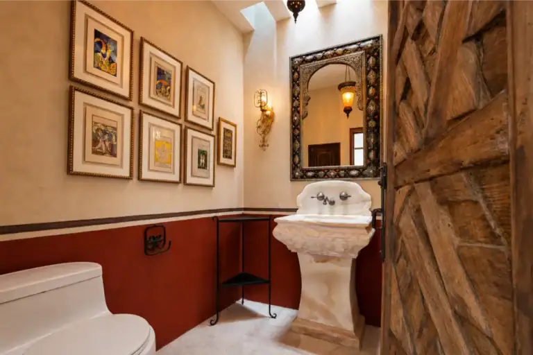 Rustic bathroom with a white pedestal sink, toilet, and a gallery wall of framed art on warm beige walls, San Miguel de Allende