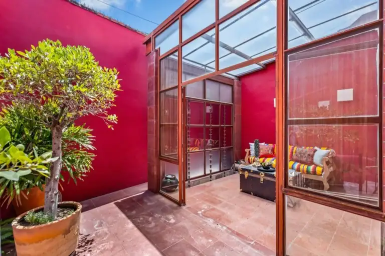 Cozy sunlit indoor courtyard with red walls, glass-framed doors, potted tree, and a seating area with colorful striped cushions around a trunk-style chest, San Miguel de Allende