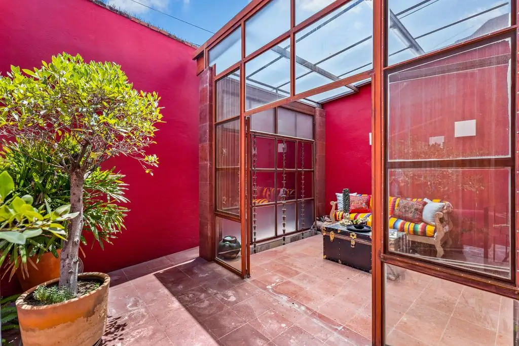 Cozy sunlit indoor courtyard with red walls, glass-framed doors, potted tree, and a seating area with colorful striped cushions around a trunk-style chest, San Miguel de Allende