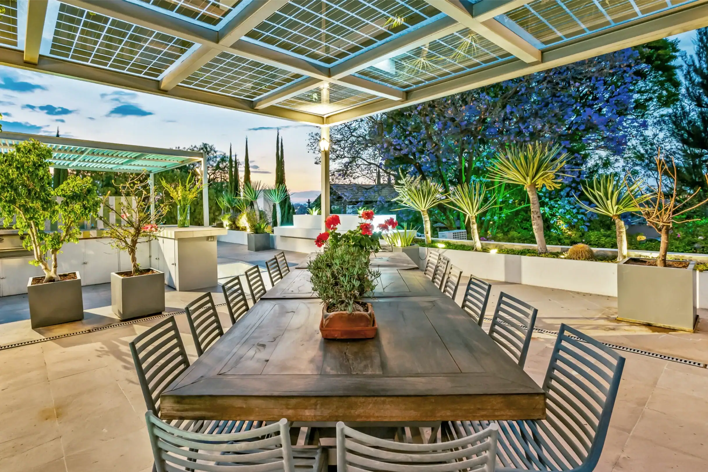 Outdoor dining setup with a long wooden table and metal chairs under a lattice pergola, surrounded by potted plants and greenery at dusk, San Miguel de Allende