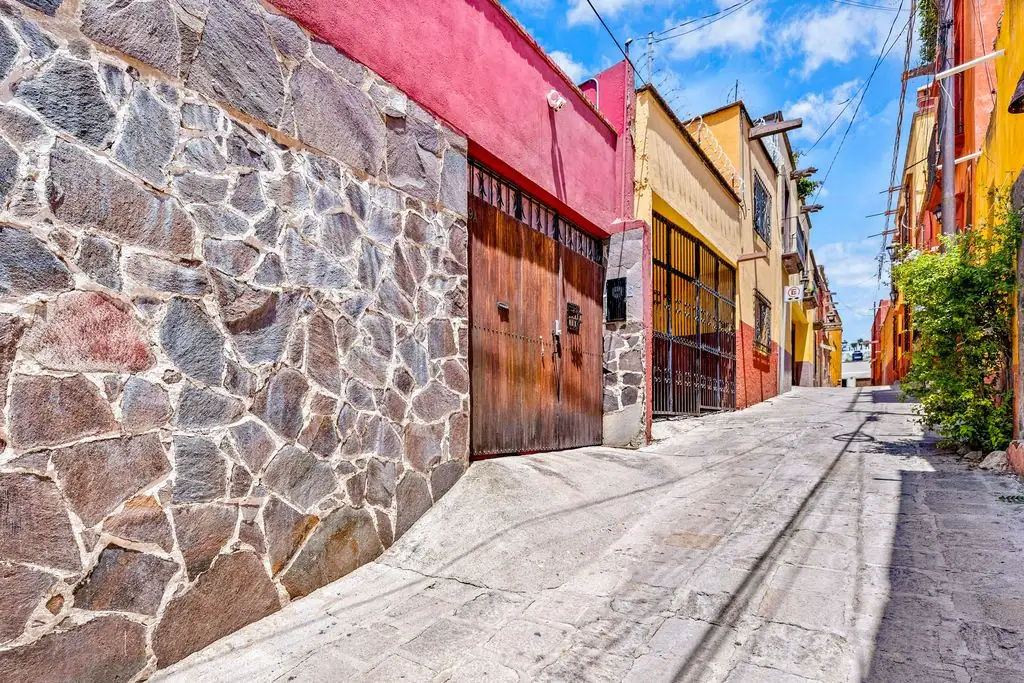 Narrow sunlit alley with a stone wall on the left and colorful buildings on the right under a blue sky, San Miguel de Allende