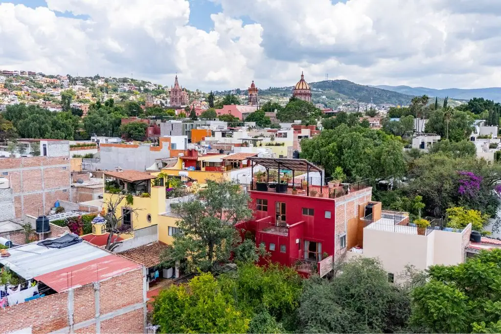Colorful hillside town with red building in foreground and cathedral domes in the distance under a cloudy skypack, San Miguel de Allende