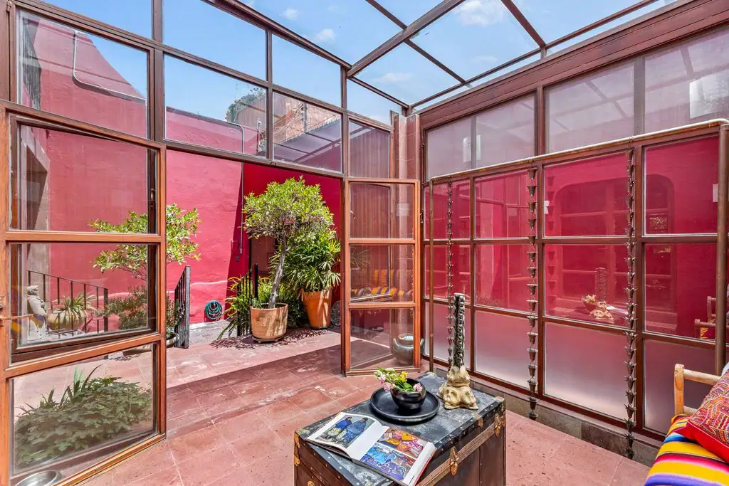 Sunlit glass conservatory with red walls, potted plants, and a wooden trunk-style coffee table with magazines on top, San Miguel de Allende