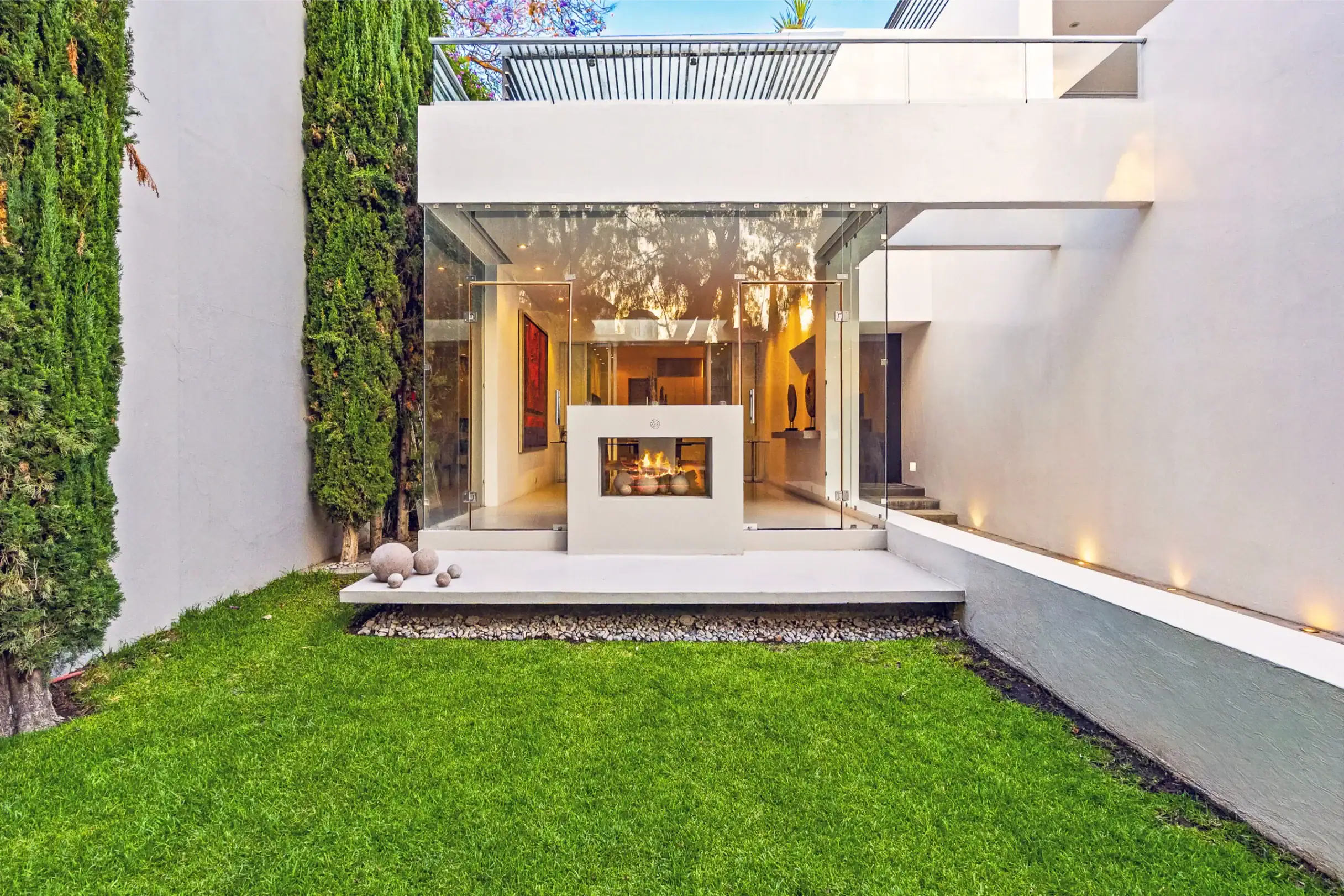Modern white house with a glass entryway, indoor fireplace visible through the doors, and a green lawn in front, San Miguel de Allende