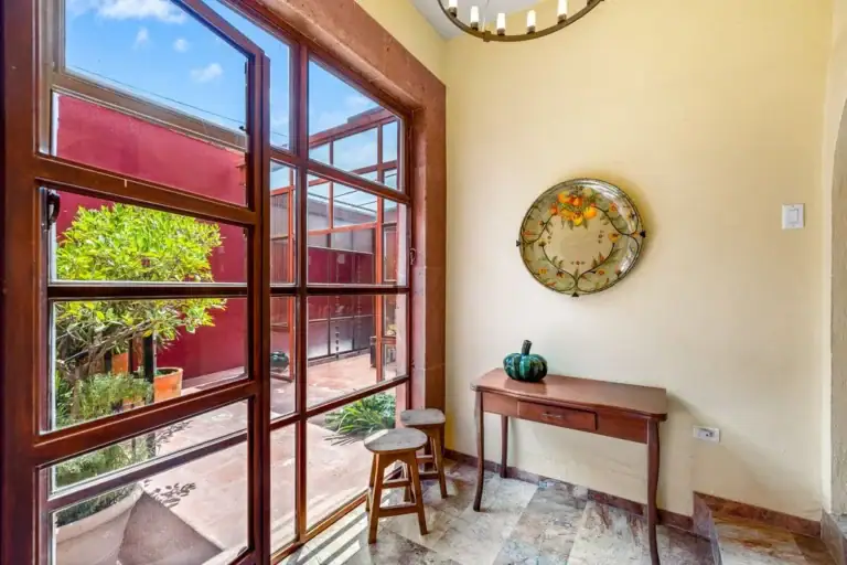 Entryway with warm yellow walls, a wooden console table, and a decorative plate; large glass doors open to a sunny courtyard with red walls and greenery, San Miguel de Allende