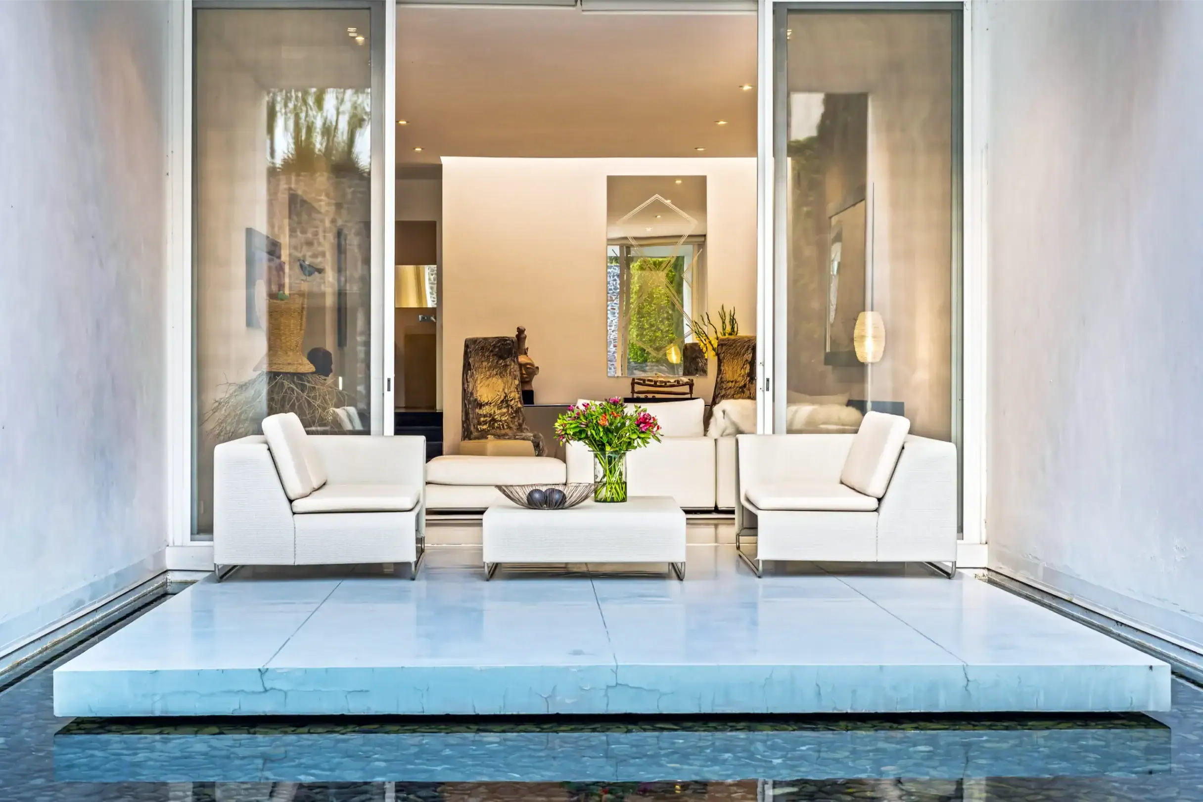 Modern outdoor living room with white cushioned seating around a glass coffee table, opening to an interior space with a blue-tiled pool in the foreground, San Miguel de Allende