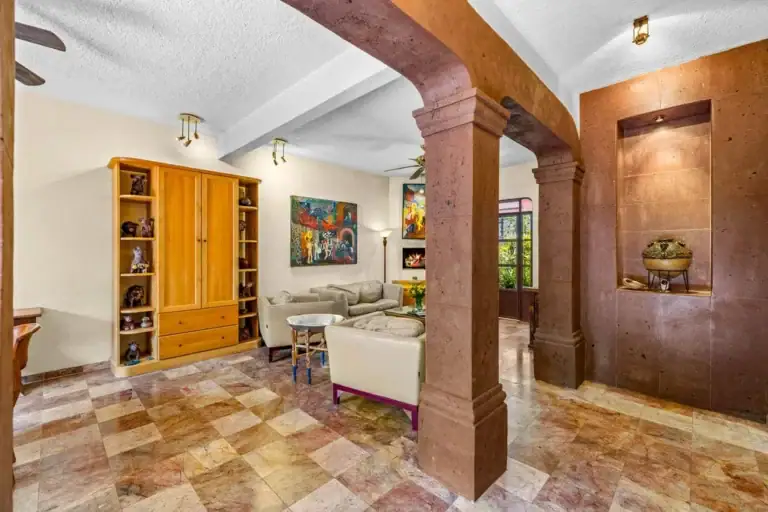 Living room with two large brown stone columns, tiled checkered floor, and beige seating area with a wooden cabinet against the wall, San Miguel de Allende