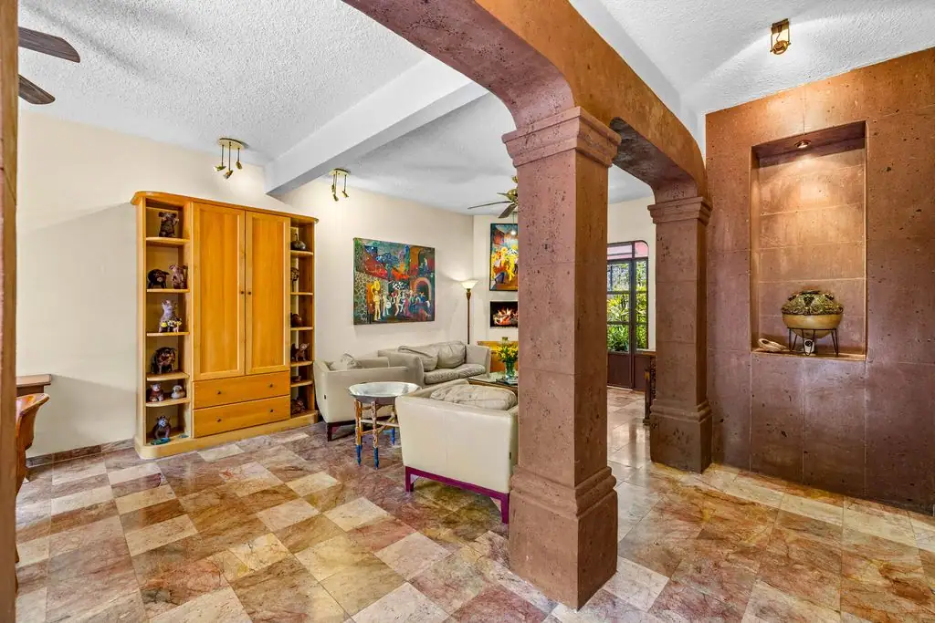 Living room with two large brown stone columns, tiled checkered floor, and beige seating area with a wooden cabinet against the wall, San Miguel de Allende