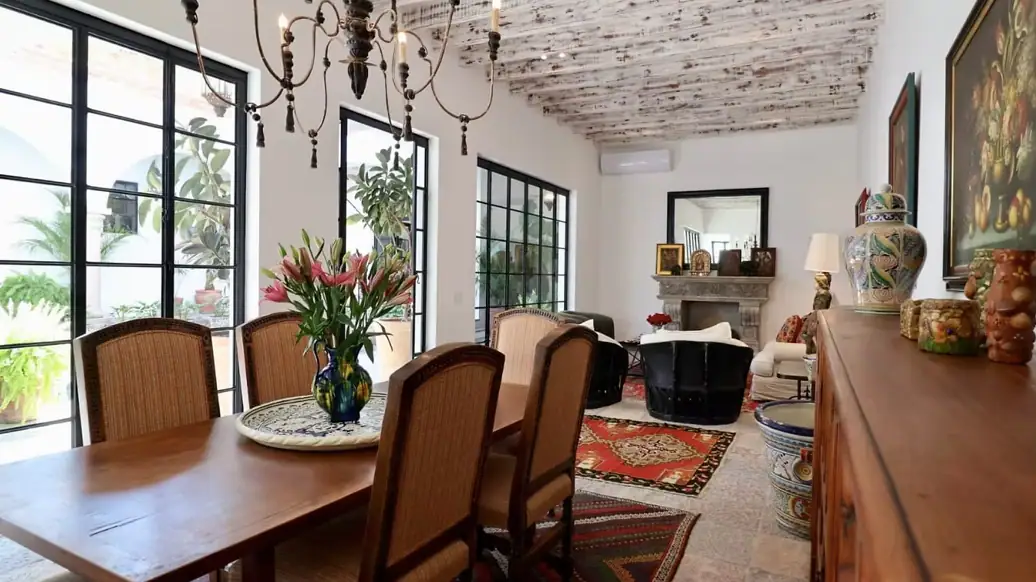 Bright dining area with a wooden table and six cane chairs, vase of pink lilies on a decorative plate, near large black-framed windows, San Miguel de Allende