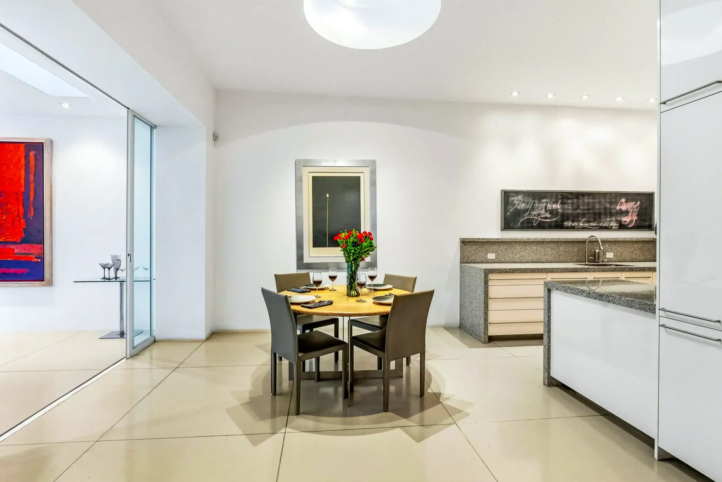Modern dining area with a round wooden table set for six, wine glasses and a vase of red flowers, in a bright open kitchen with light cabinetry and a chalkboard wall, San Miguel de Allende
