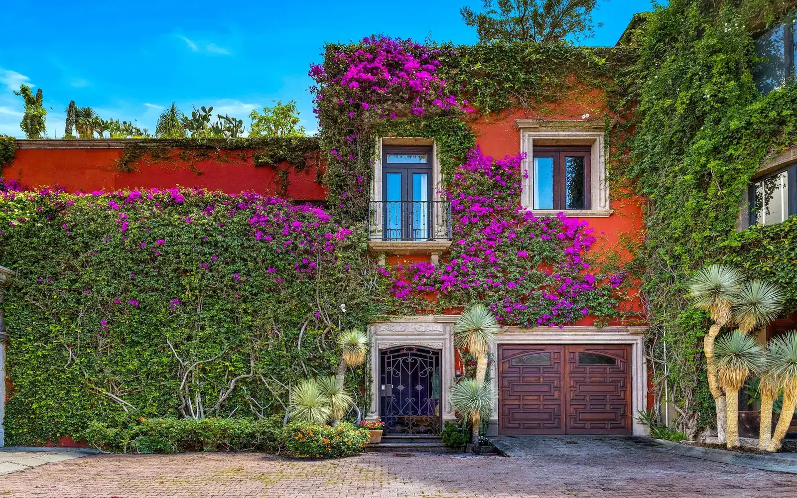 Red stucco building facade covered in green ivy and magenta bougainvillea, with two windows and a small balcony, San Miguel de Allende