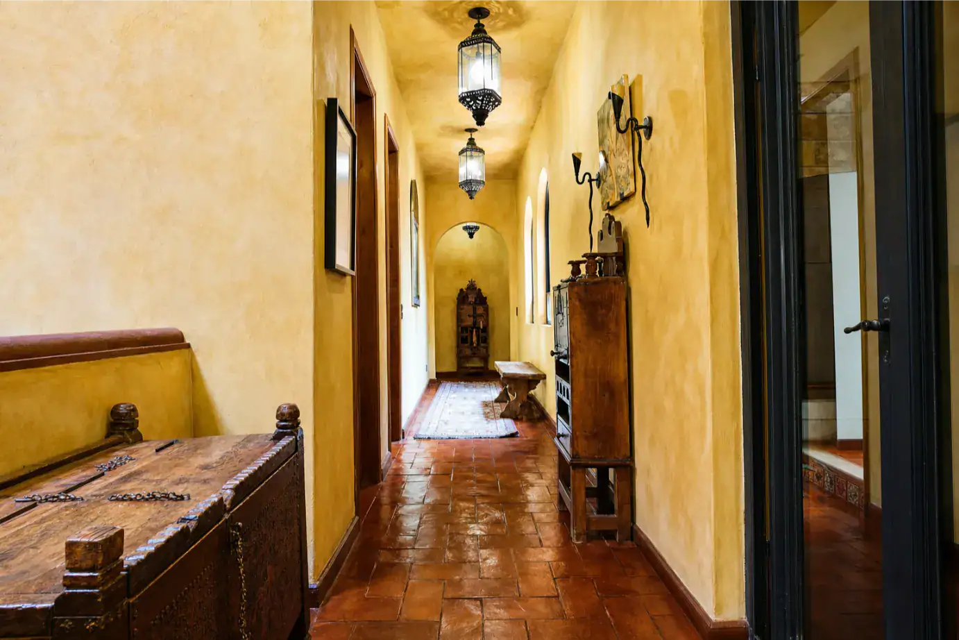 Warm yellow hallway in a historic home with antique wooden chest, benches, and hanging lanterns, San Miguel de Allende