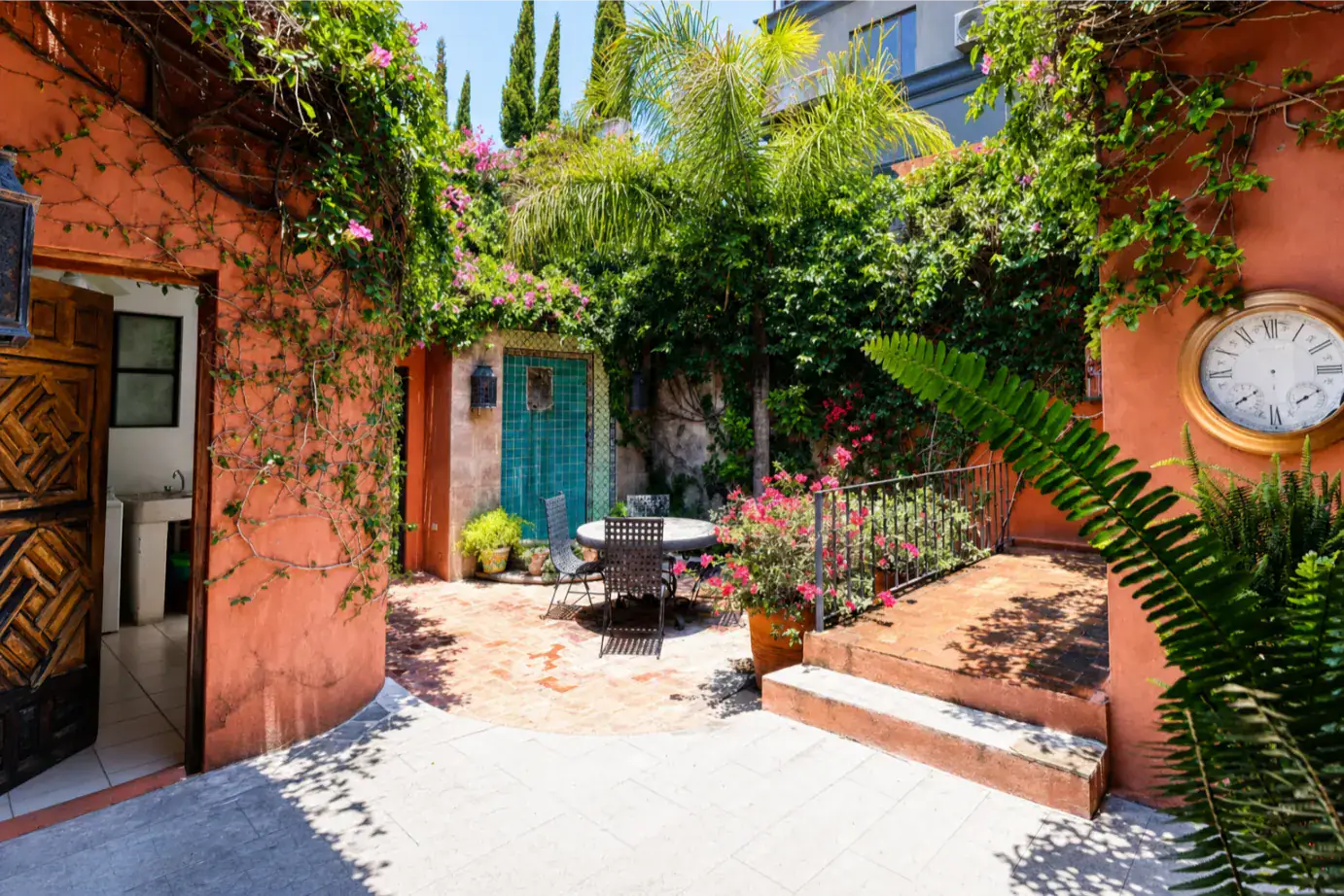 Terracotta-walled courtyard with climbing vines, potted plants, and a round table with chairs under a sunny sky, a large clock on the right wall, San Miguel de Allende