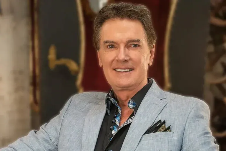 Greg Gunter smiling in a light blue-gray blazer over a dark patterned shirt with a dark pocket square, seated in front of an ornate interior with red velvet and gilded details