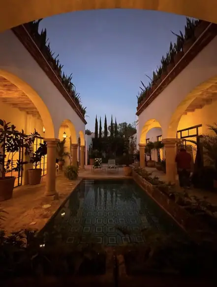 Archways surround a narrow rectangular pool in a courtyard at dusk, with warm amber lighting, potted plants, and seating in the background, San Miguel de Allende