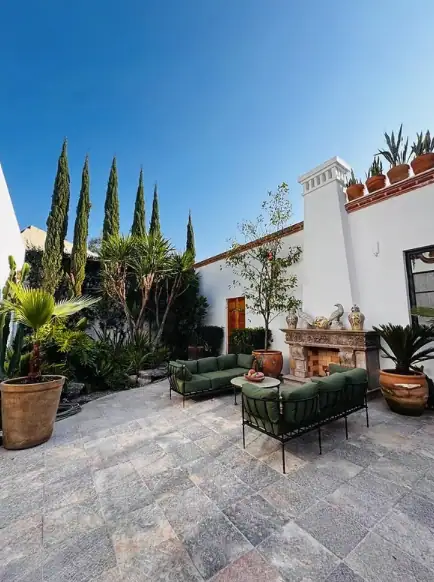 Sunny outdoor patio with white walls, tall cypress trees, and a stone tile floor. Green cushioned seating around a stone fireplace with potted plants nearby, San Miguel de Allende